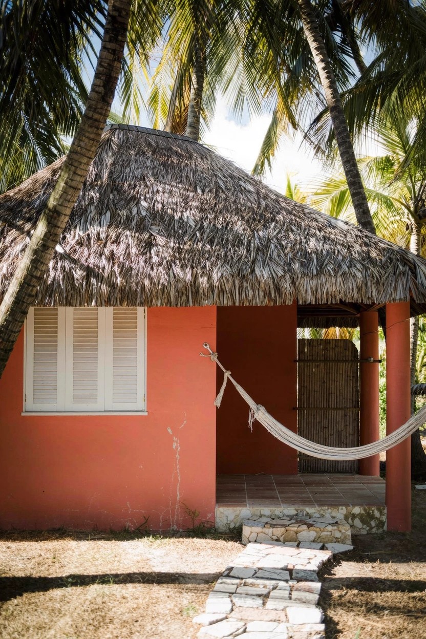 Pink bungalow-style house with thatched roof overhanging a porch with hammock, wooden shutters and door, stone pathway, palm trees, and dry ground.
