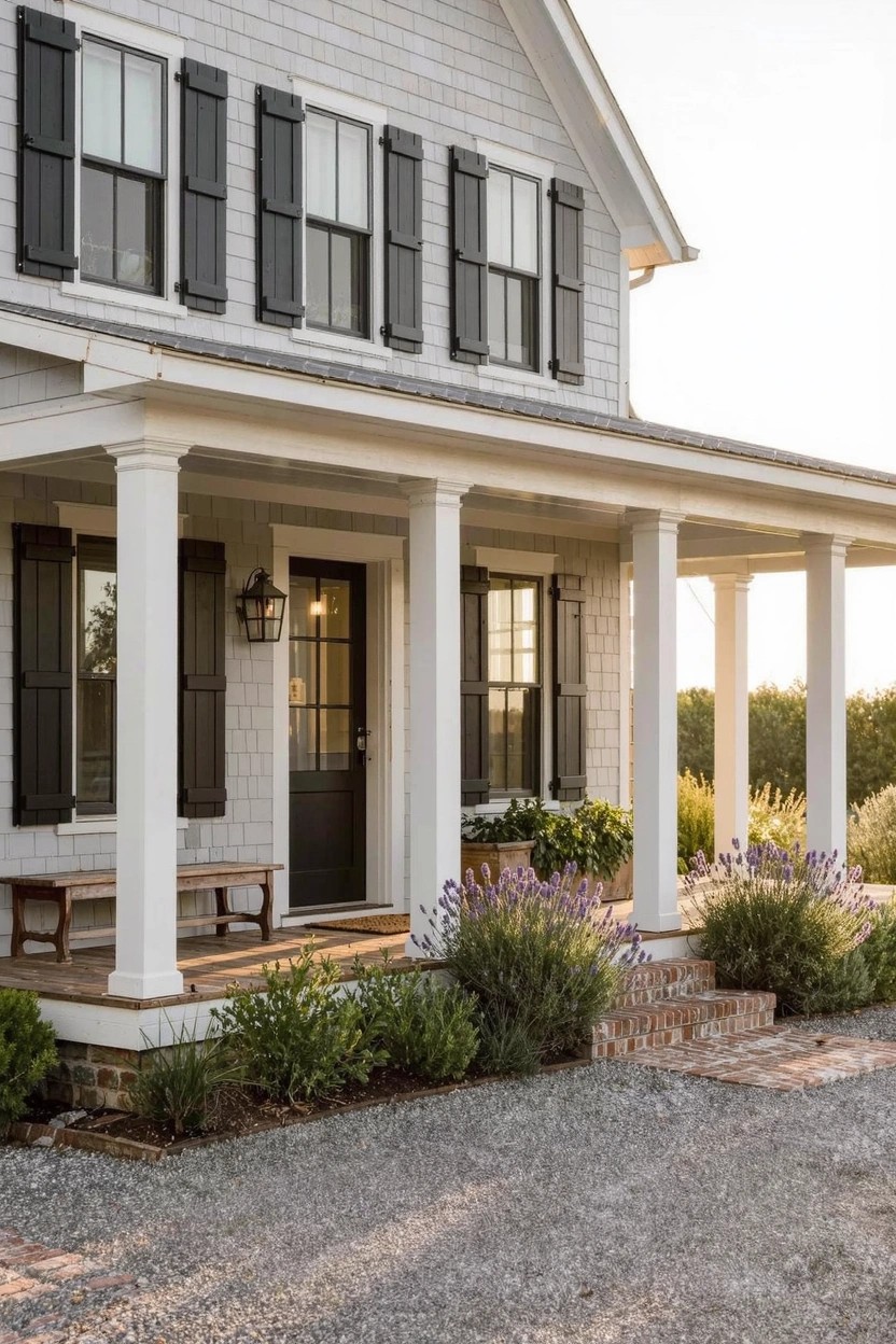 Light gray clapboard house exterior with black shutters and door, white columns supporting a covered porch, hanging lanterns, potted plants, and gravel driveway with brick steps.