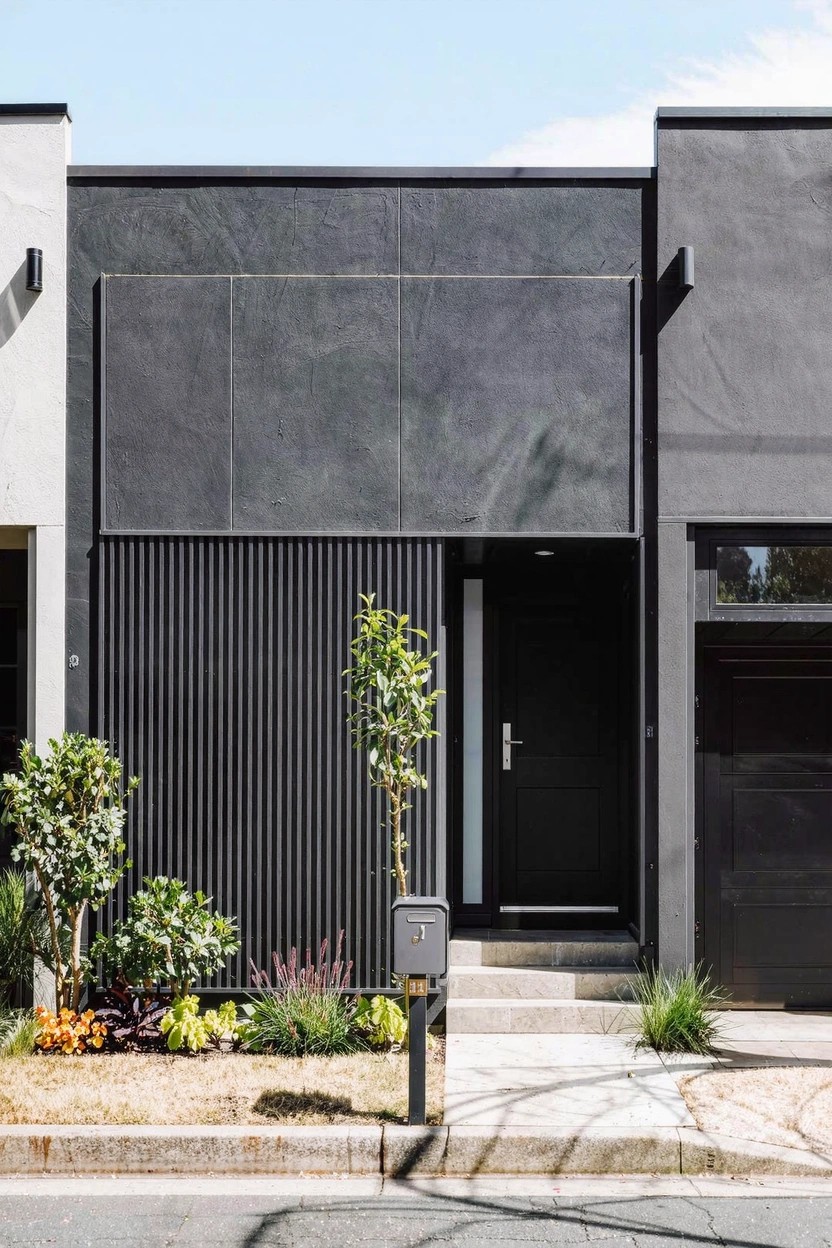 Modern house exterior featuring dark gray cladding with vertical slats beside a black front door, black garage door, concrete steps, sparse plants, gravel ground cover, and a mailbox in the front yard.