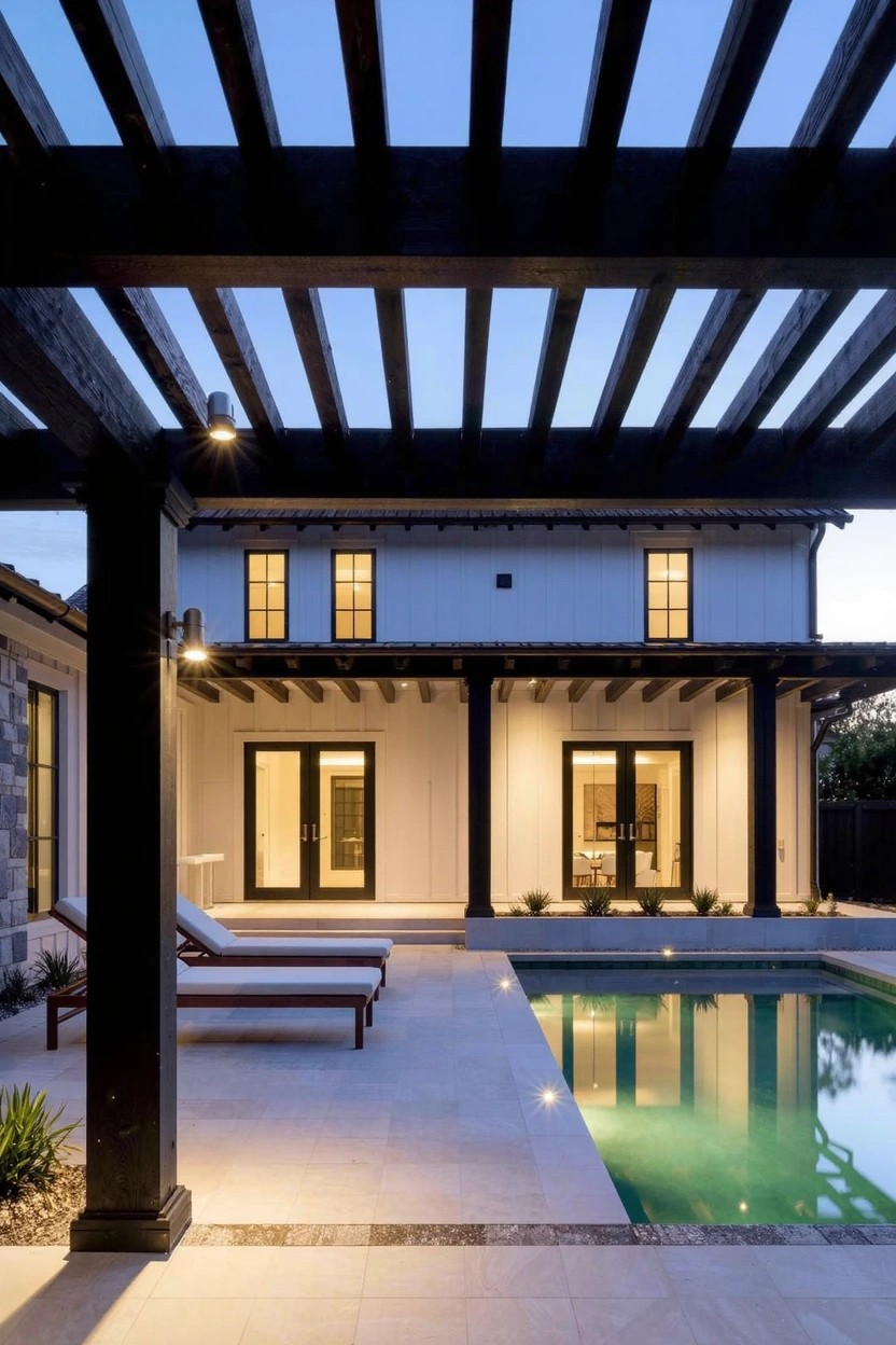 Backyard view of white stucco house with black wooden pergola over concrete pool deck and lounge chairs, pool lit at dusk with wall and post lights.