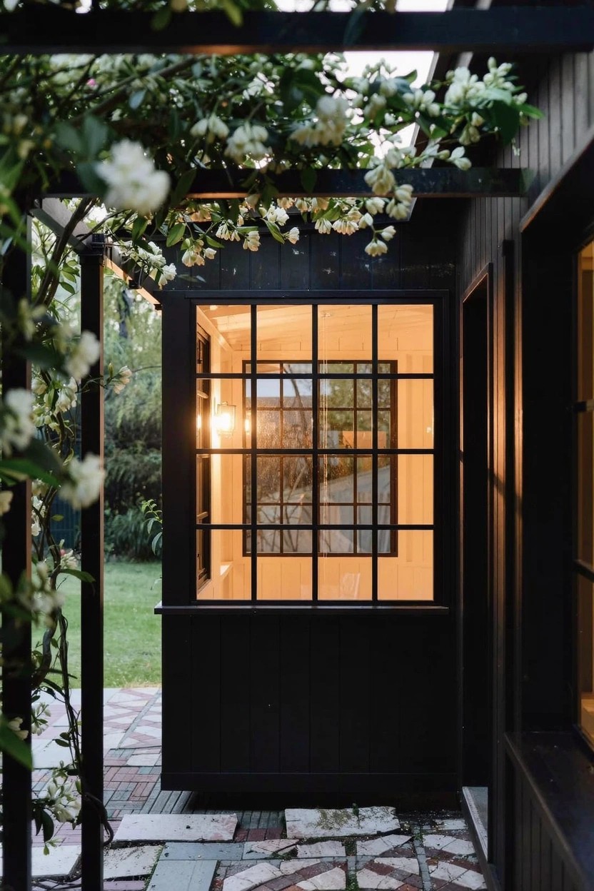 Black wooden pergola overgrown with white flowering vines arches over a brick-paver path leading to a large grid window on a dark exterior wall, with green lawn and plants nearby.