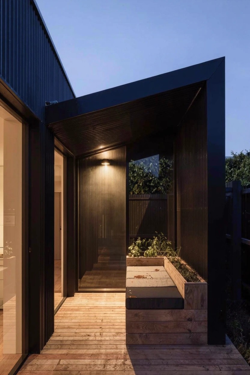 Modern house exterior at dusk with black corrugated metal walls and cantilevered roof over a wooden deck, featuring a built-in bench planter, glass sliding doors, wall light, and nearby greenery.