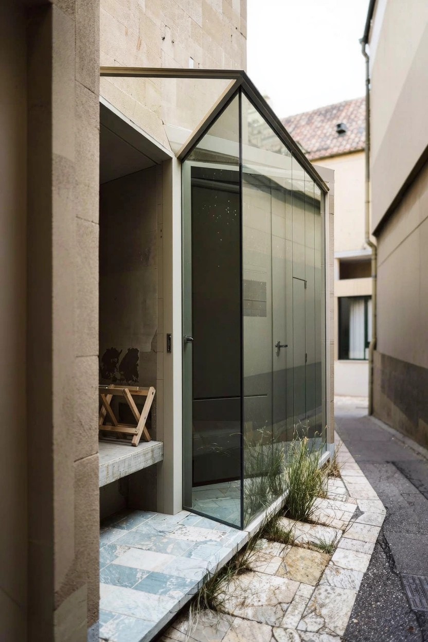 Narrow exterior alley view of a beige stone house with a protruding glass-enclosed entry structure containing a wooden chair, adjacent stone walls, tiled step, and sparse plants.