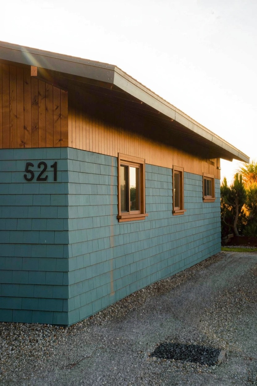 Side view of a small building with light blue shingle siding on lower walls, brown horizontal wood siding above, three rectangular windows, black number 521 on the wall, gravel driveway, and distant plants at sunset.