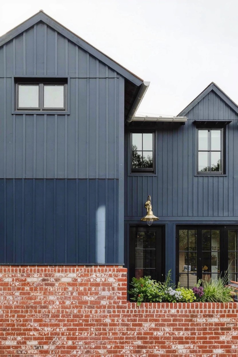 House exterior featuring navy blue vertical board-and-batten siding on the upper stories above a red brick lower wall, with large black-framed windows, a glass front door flanked by sidelights, a gold lantern light, and greenery along the brick base.