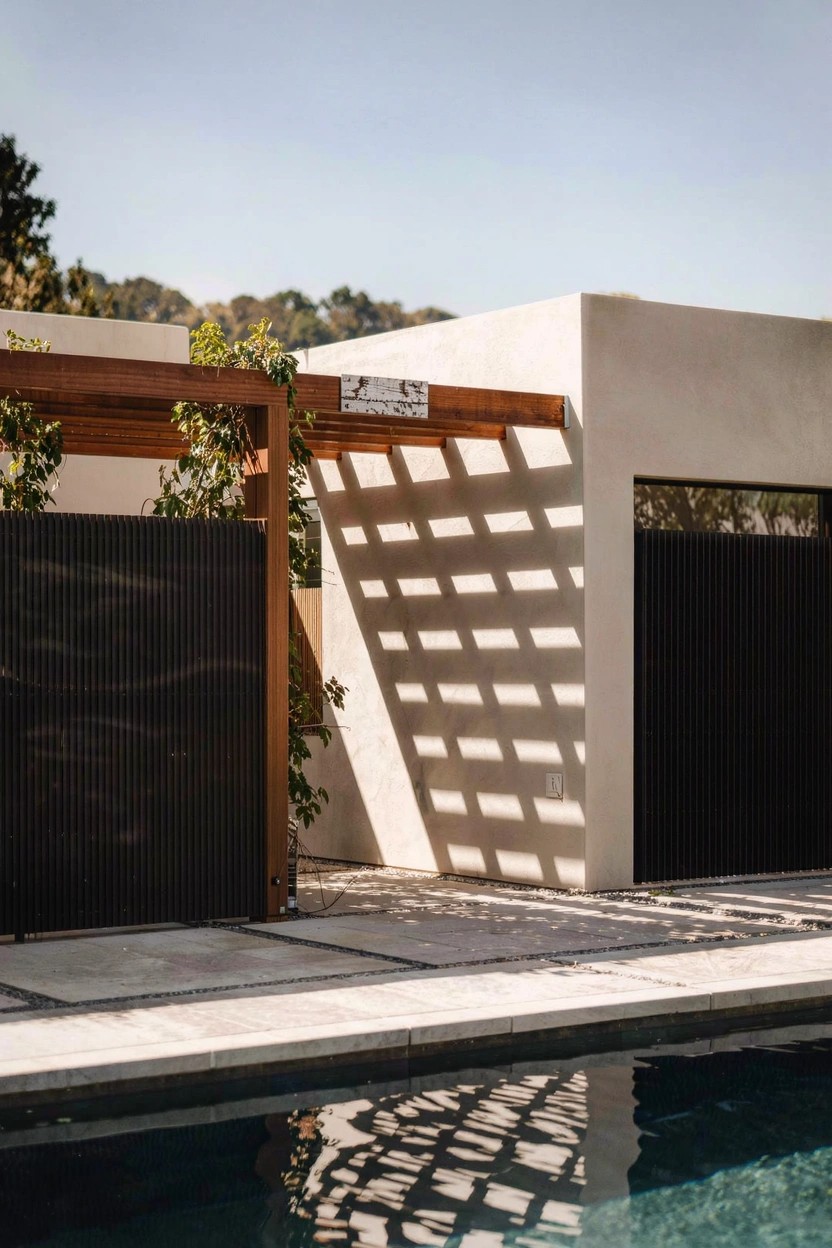 Beige stucco exterior wall next to dark vertical slatted wood gates, wooden lattice pergola overhead with vines, stone pavers on ground, and swimming pool reflecting shadows in sunlight.
