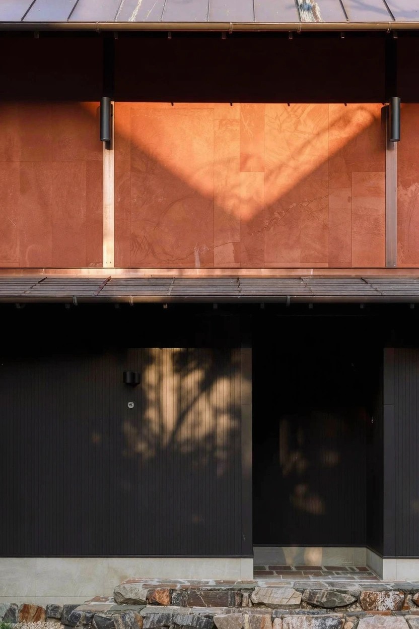 House exterior with reddish plank wood siding on upper walls under dark metal roof overhang, dark vertical wood siding on lower entry area, and stone retaining wall at base.