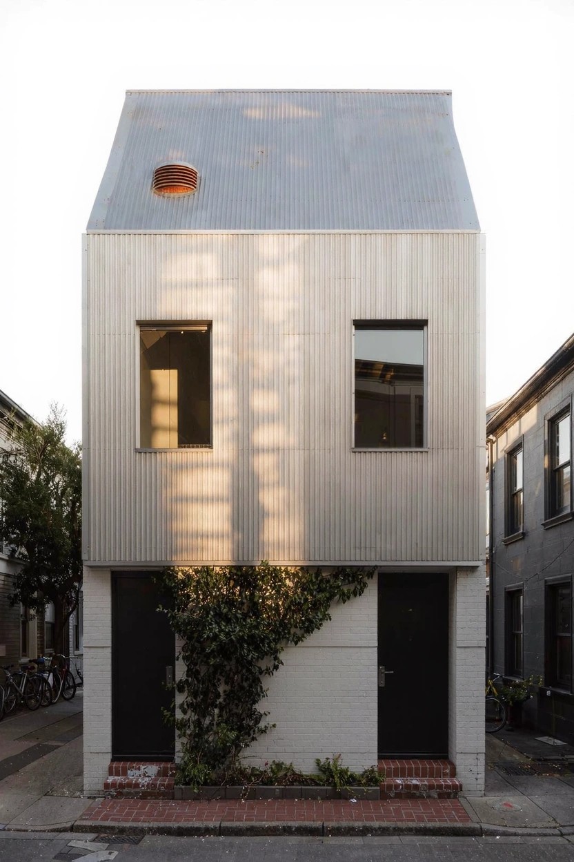 A narrow two-story house with gray vertical corrugated metal siding, two black doors on the ground floor with ivy climbing beside them, rectangular windows, and a gabled metal roof with a small vent, between brick buildings on a street.