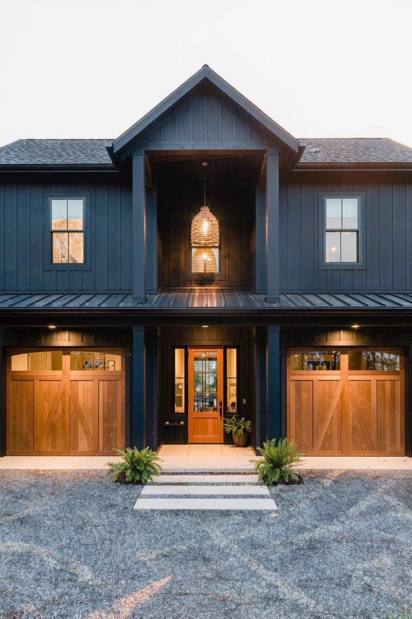 Two-story modern farmhouse house exterior with dark navy board-and-batten siding, black metal roof, wooden garage doors, central front entry with glass door and rattan pendant light, potted plants flanking concrete steps, gravel driveway.
