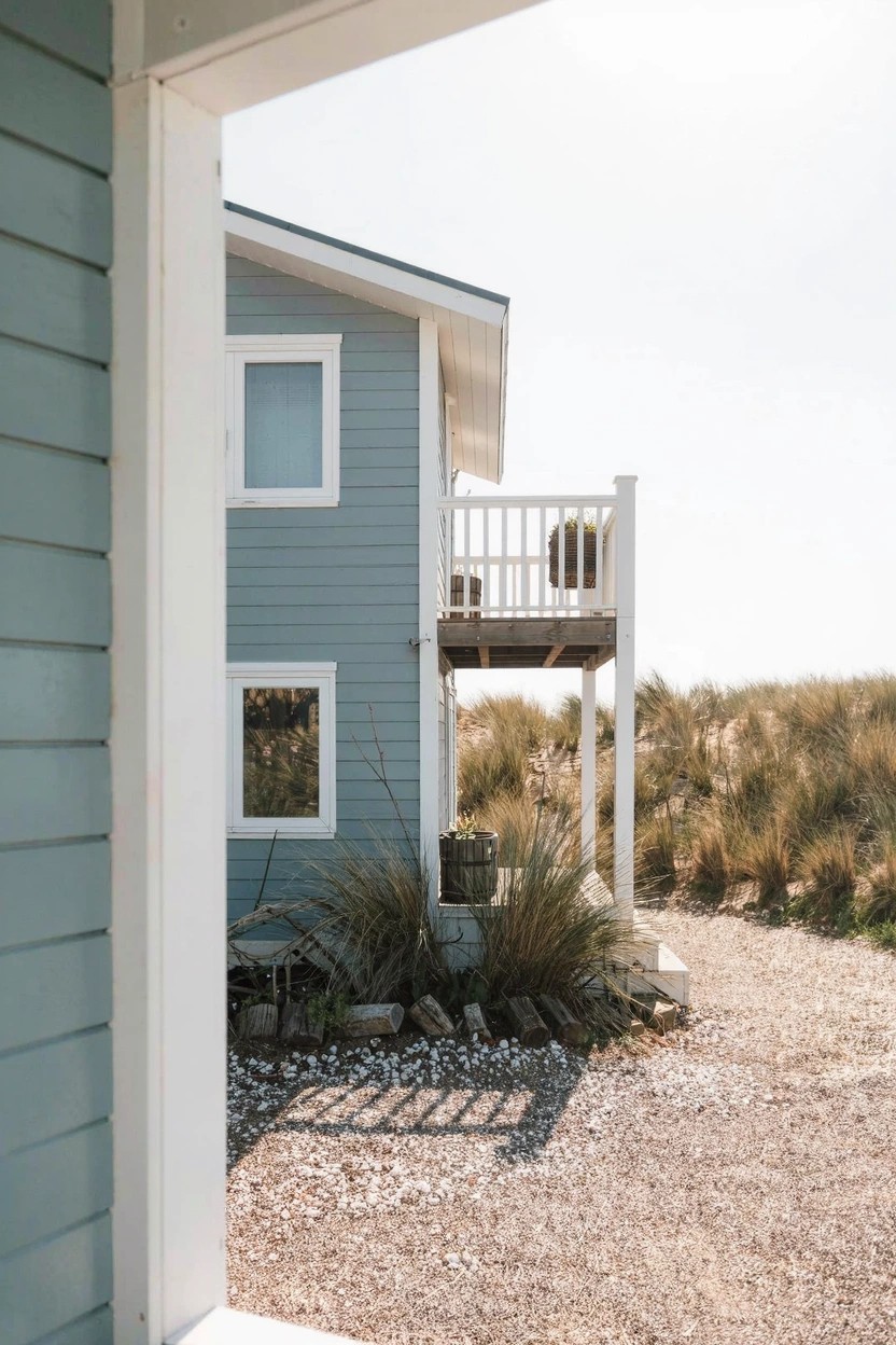 Side view of a light blue clapboard house with white window frames, a second-story balcony with white railing, dune grasses, shrubs, and a gravel path.