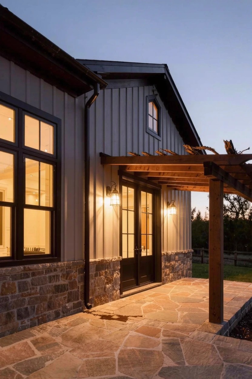 House exterior at dusk with light gray corrugated metal siding, beige stone base, black windows and door under a wooden pergola, stone paver walkway, and wall lanterns.