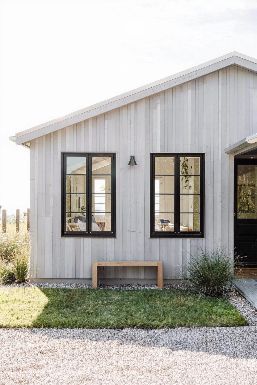 White board-and-batten house exterior with two black-framed windows, wall light, wooden bench on gravel path, ornamental grasses, and green lawn.
