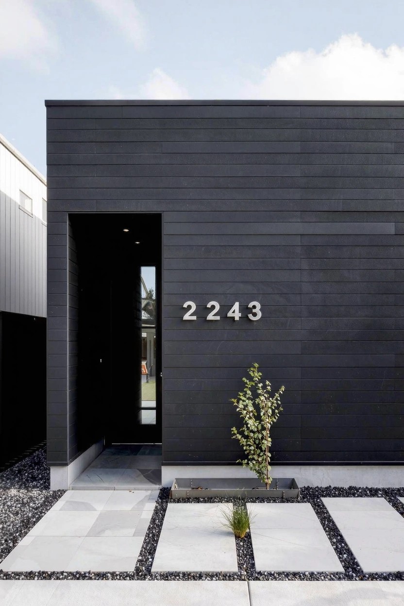 Modern house exterior featuring vertical black cladding, recessed entry with glass door and white house number 2243, light paver pathway, gravel ground cover, and small plants.