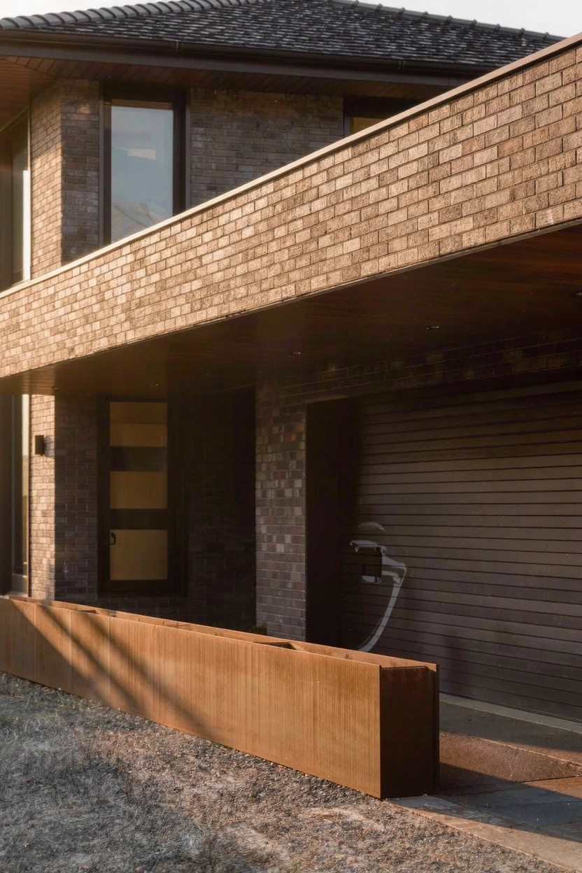 Brick house exterior featuring a dark garage door, overhanging upper level, rusted metal planter box along the front edge, gravel driveway, and EV charger cable.