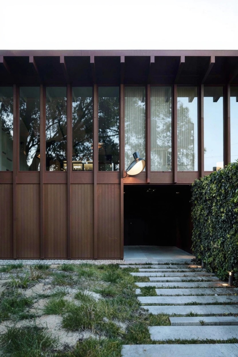 Modern single-story house exterior featuring vertical dark wood cladding, large horizontal glass windows with sheer curtains, a large white hanging light fixture over a recessed entrance, concrete steps pathway, hedges, and sparse grass landscaping.