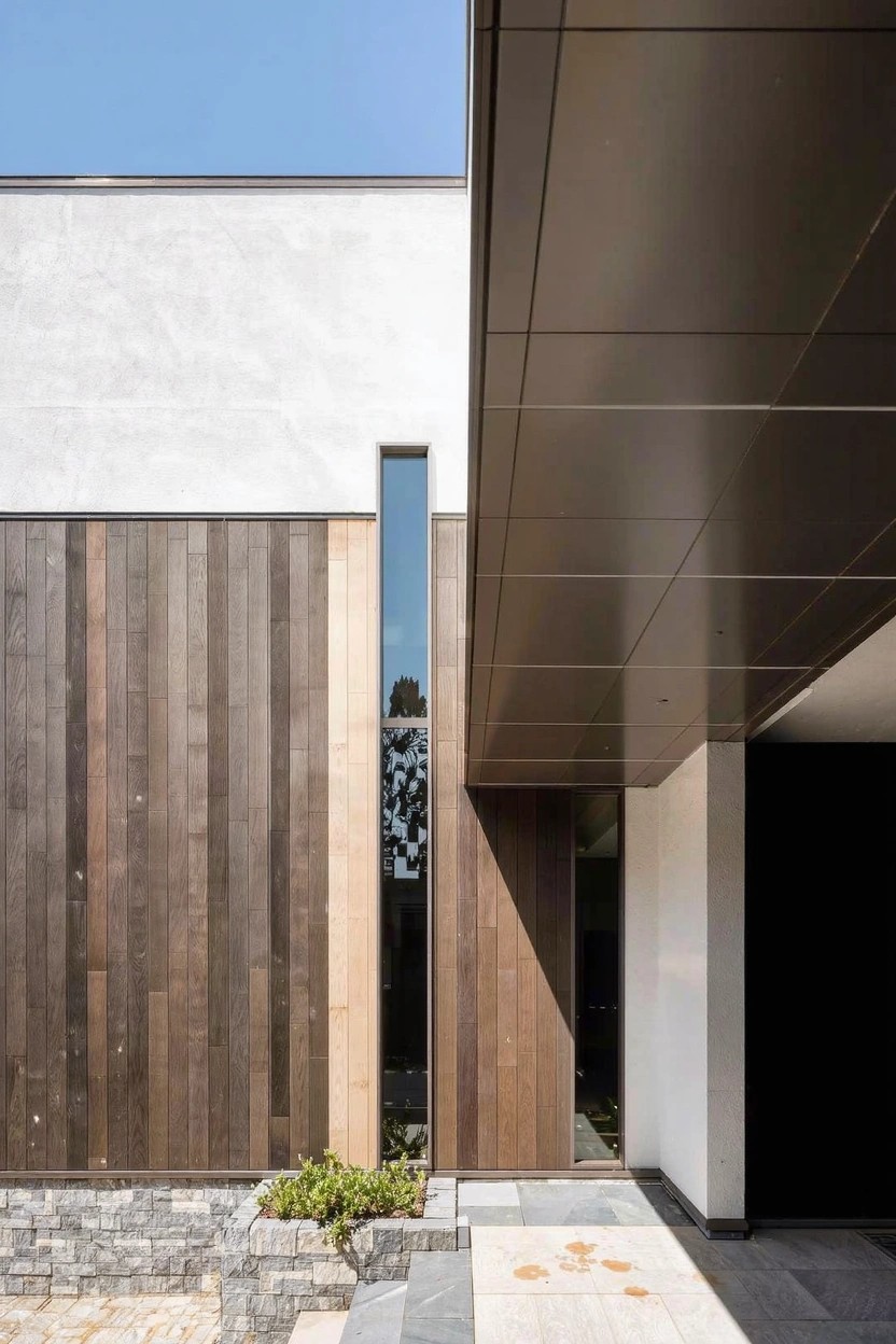 Modern small house exterior with white plaster walls, vertical warm-toned wood cladding on one side, dark metal overhanging canopy above a wood entry door, narrow vertical window, stone base and pathway, and sparse greenery.