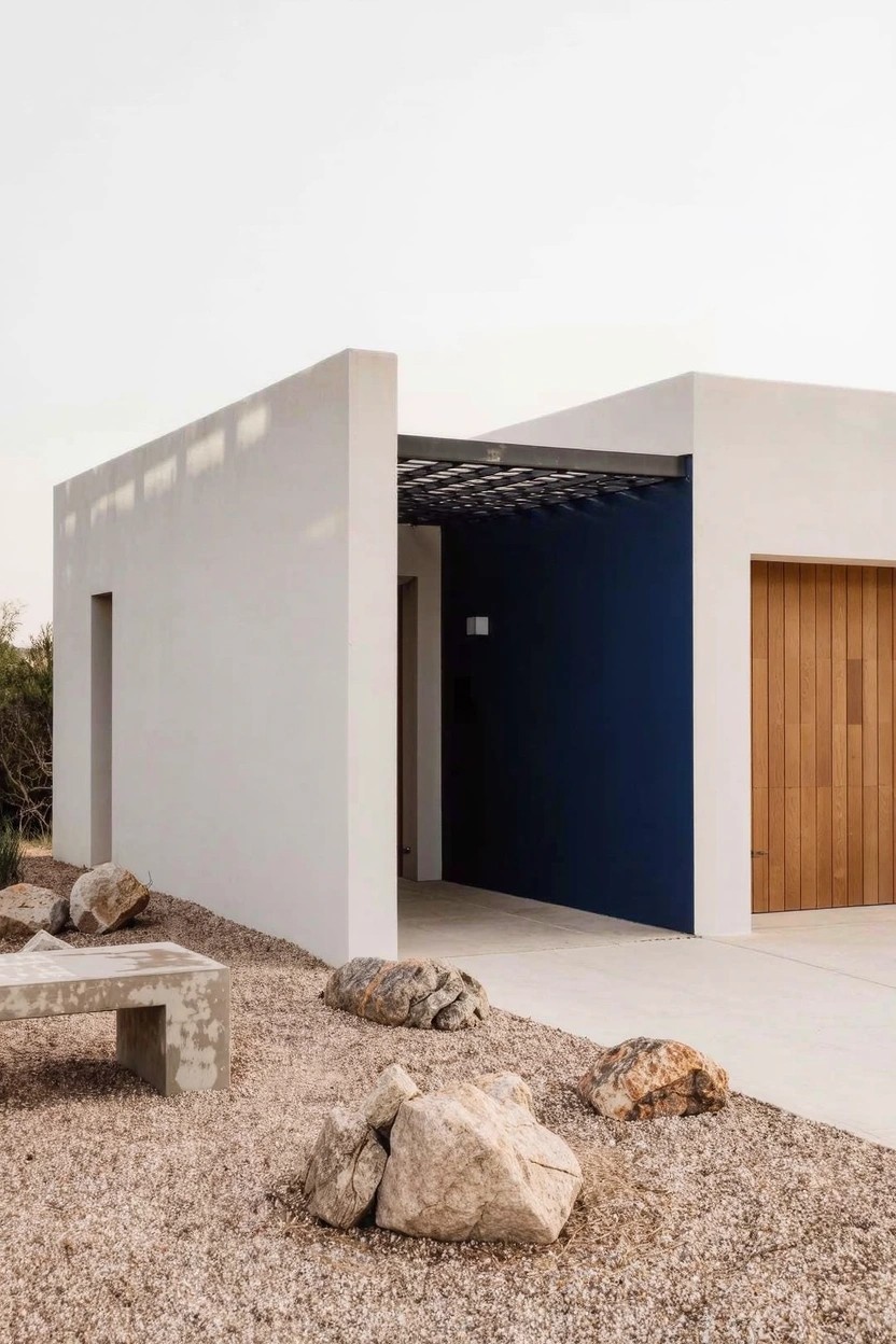 Modern house exterior featuring white stucco walls, a deep blue accent wall beside a wooden garage door, covered entry with lattice overhead, concrete bench, gravel ground, and scattered large rocks.