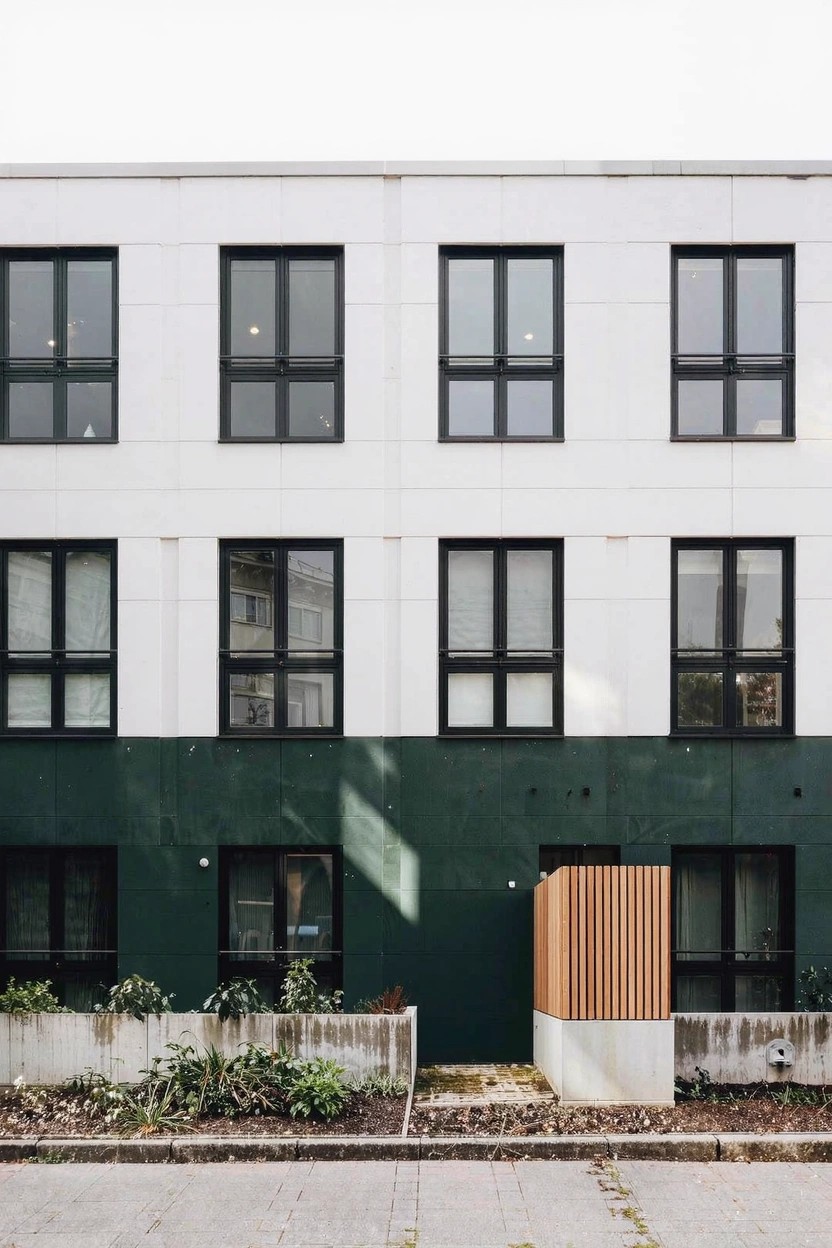 Multi-story modern building exterior with white upper walls and large dark-framed windows above a dark green lower section, wooden slatted entry feature, concrete planters with plants, and a paved pathway.