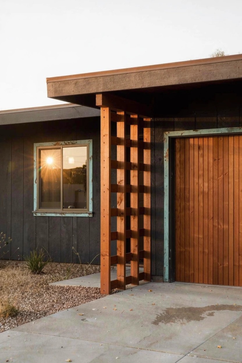 Contemporary house exterior with dark gray siding, turquoise-framed window, wooden garage door, tall reddish wooden posts with horizontal lattice screening, concrete driveway, sparse desert plants, and sunset sky.