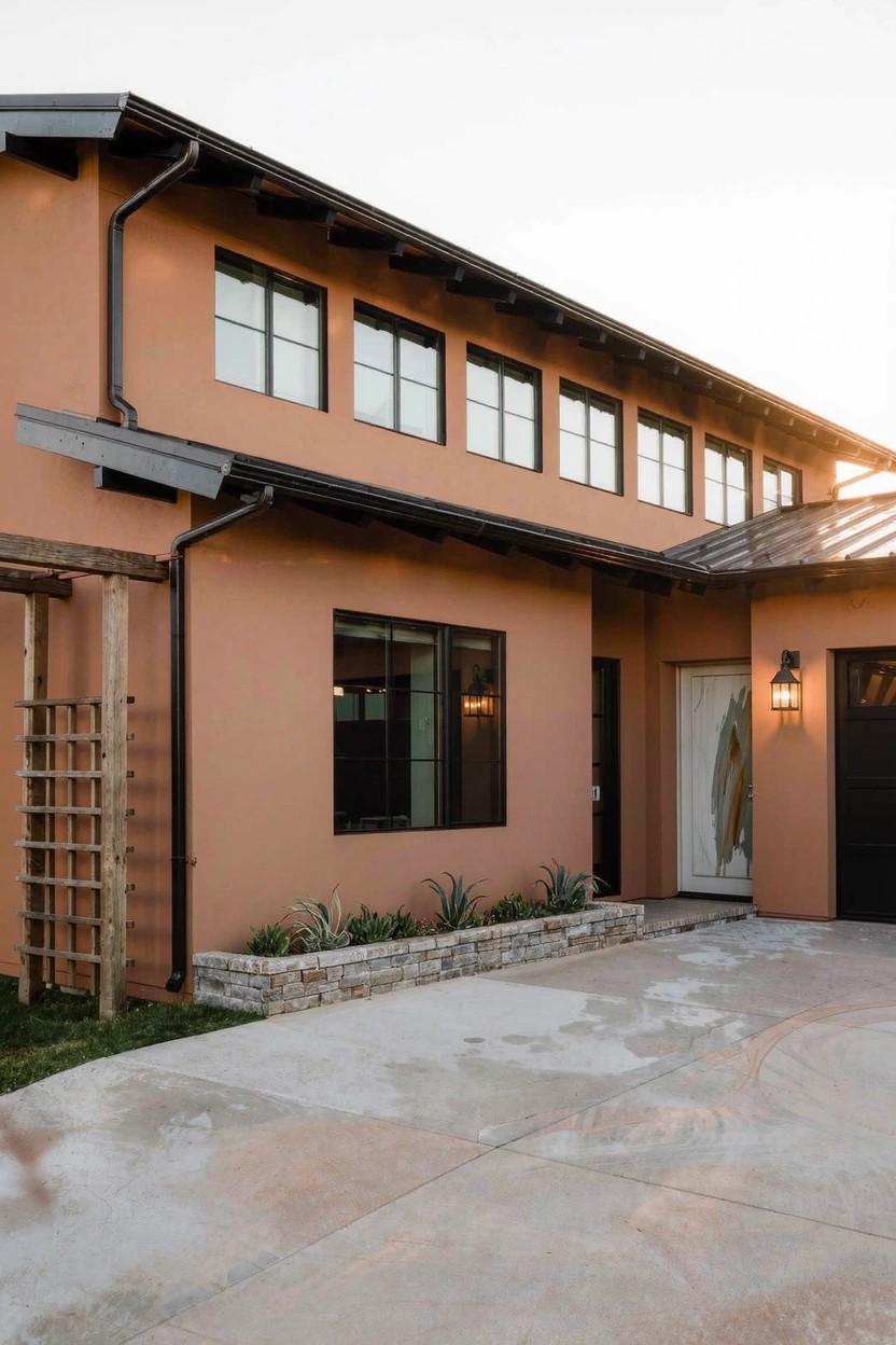 Contemporary house exterior featuring terracotta stucco walls, black-framed windows, dark metal roof, wooden pergola with plants, white entry door, and concrete driveway.