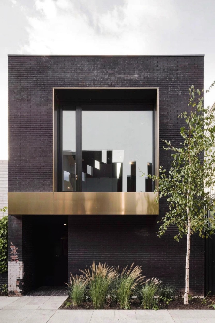 Two-story modern house with black brick facade, large vertical window framed in brass metal, brass surround at recessed entry, tall ornamental grasses and birch tree at front, paved walkway.