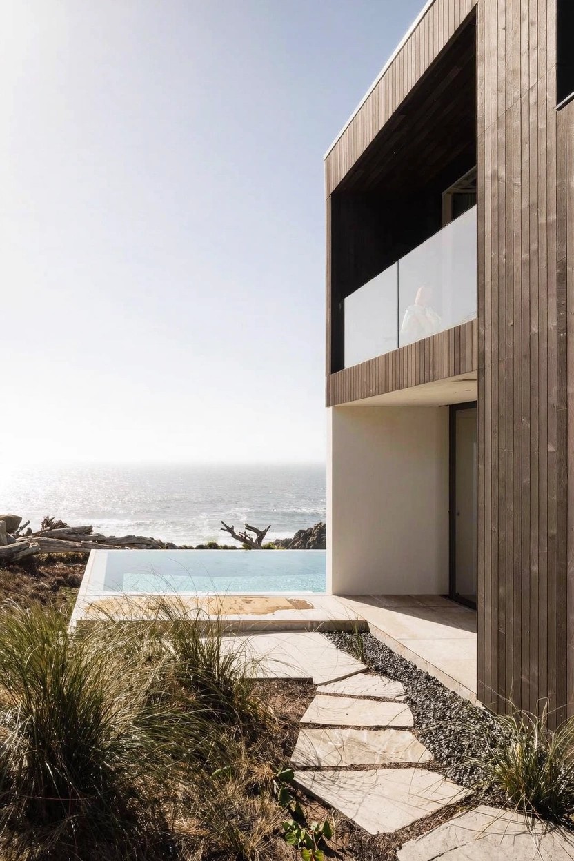 Modern two-story house exterior featuring dark wood cladding on upper sections, white walls below, an infinity-edge pool at the corner facing the ocean, with native grasses, a stepping stone path, and distant waves visible.