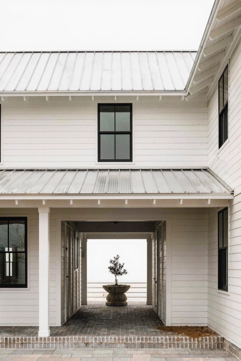 White clapboard house exterior with metal roof, black-framed windows, covered breezeway supported by columns on brick pavers, and a potted tree on a pedestal centered in the breezeway.