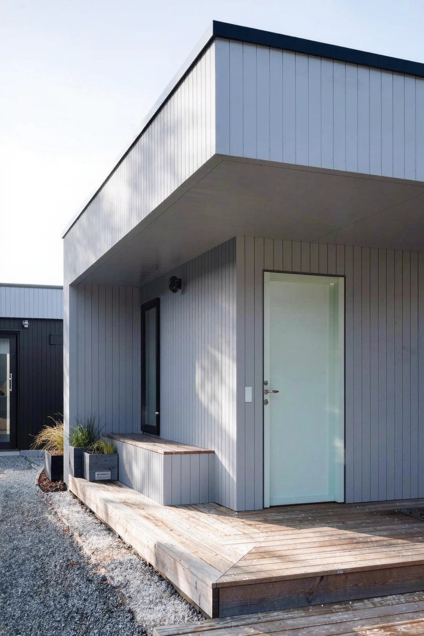 Corner view of a modern house with light gray vertical siding, white entry door under overhang, black trim, wooden deck porch, potted plants, and gravel ground beside another dark building.