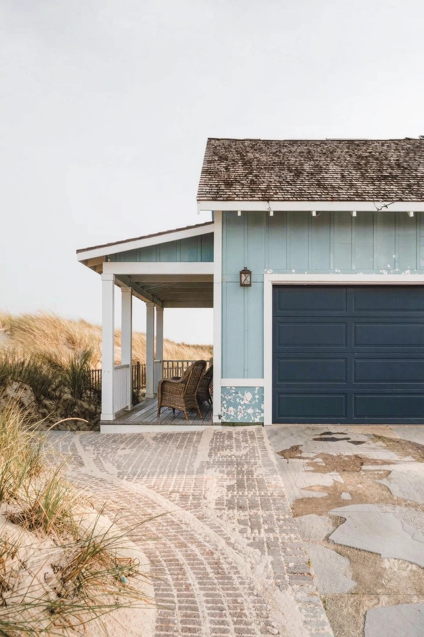 Light blue house with shakes roof, white-trimmed porch, navy garage door, brick pathway, and dune grass nearby.