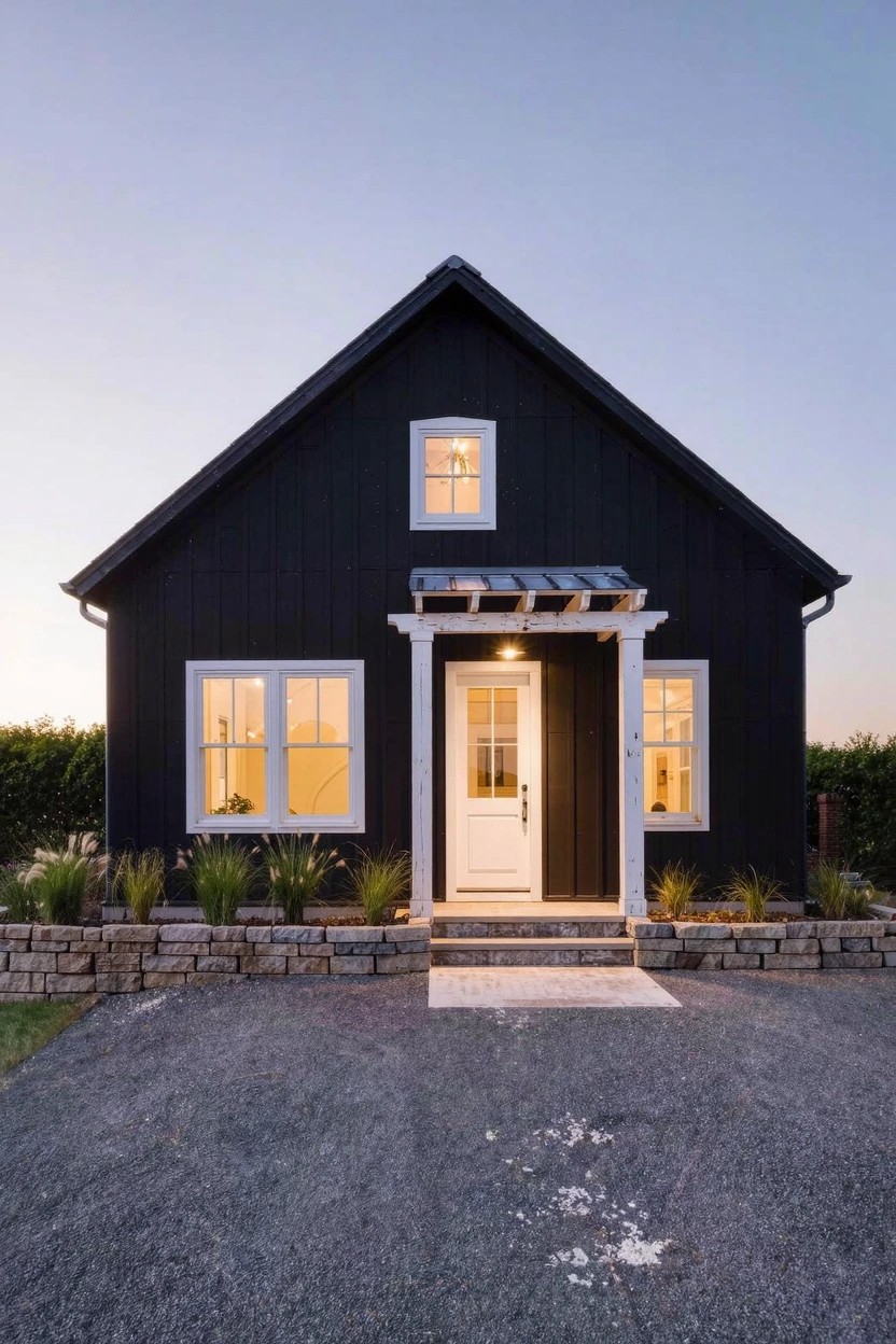 Black clapboard house with gabled metal roof, white-trimmed windows and door under a covered porch with white columns, stone retaining walls, ornamental grasses, and gravel driveway at dusk.