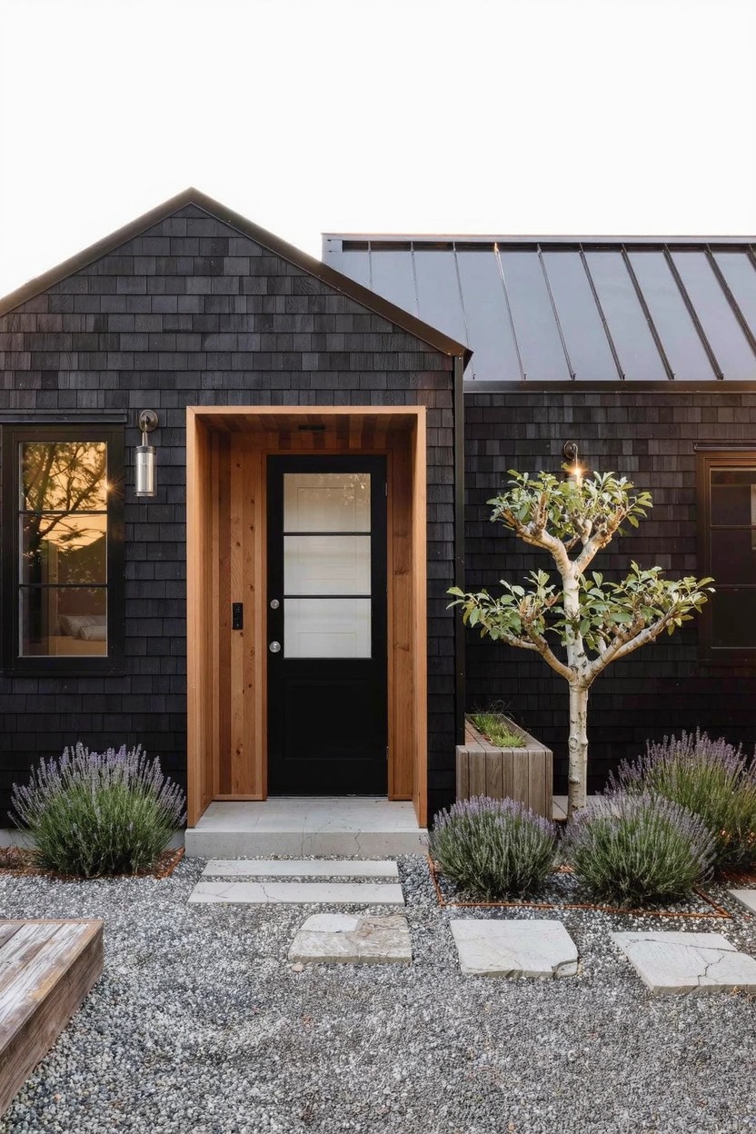 Small house exterior with black shingle siding, dark metal gable roof, recessed wood-framed glass entry door flanked by windows, black wall lantern, gravel yard, stone step path, lavender plants, wooden planter with small tree, and wooden bench.
