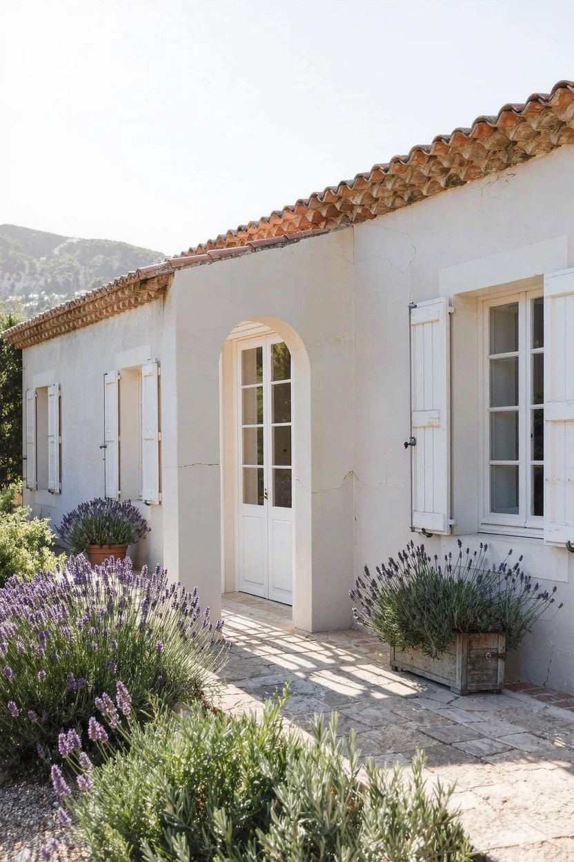 White stucco house exterior with terracotta tile roof, arched white double door entry, white shuttered windows, stone pathway lined with lavender plants, and hillside backdrop.