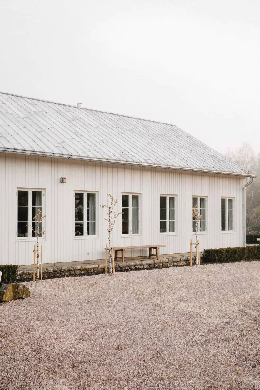 White clapboard building with gray standing seam metal roof, multiple white-framed windows along the facade, gravel ground, young trees, and wooden bench in front.