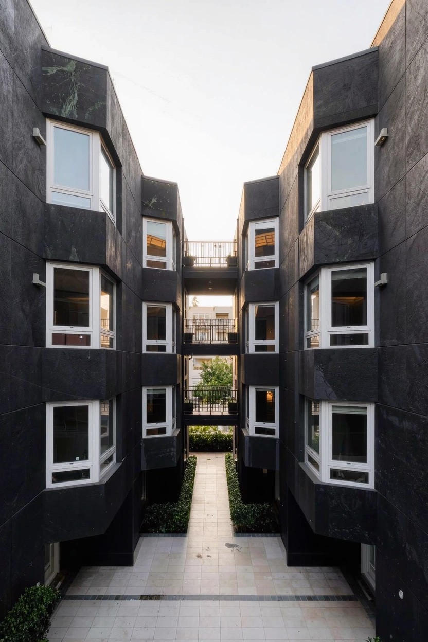 Multi-story modern building with black angular walls, numerous white-framed rectangular windows, balconies, and a central stone pathway flanked by greenery.