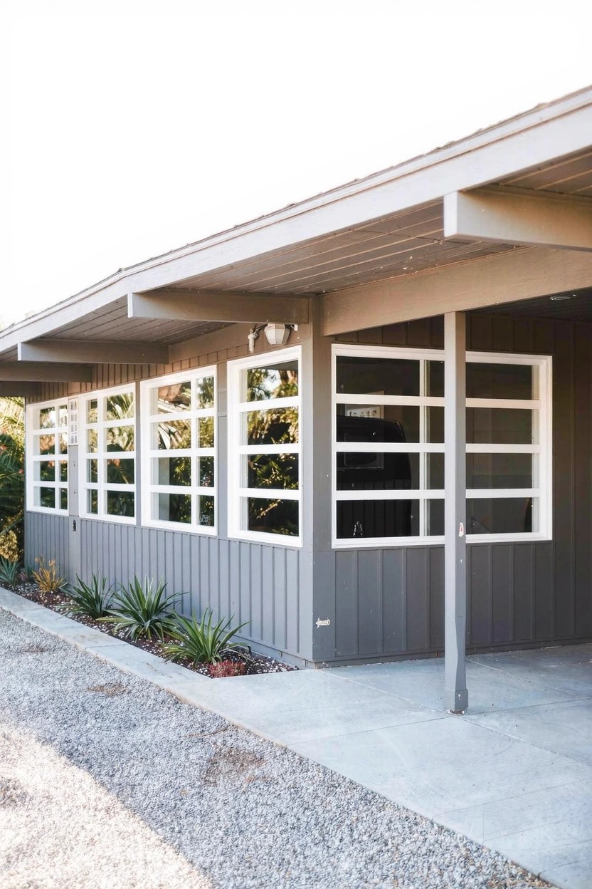 Side view of a gray wood-sided house featuring multiple large white-framed multi-pane windows under a covered overhang, with a gravel driveway, concrete entry slab, and agave plants along the base.