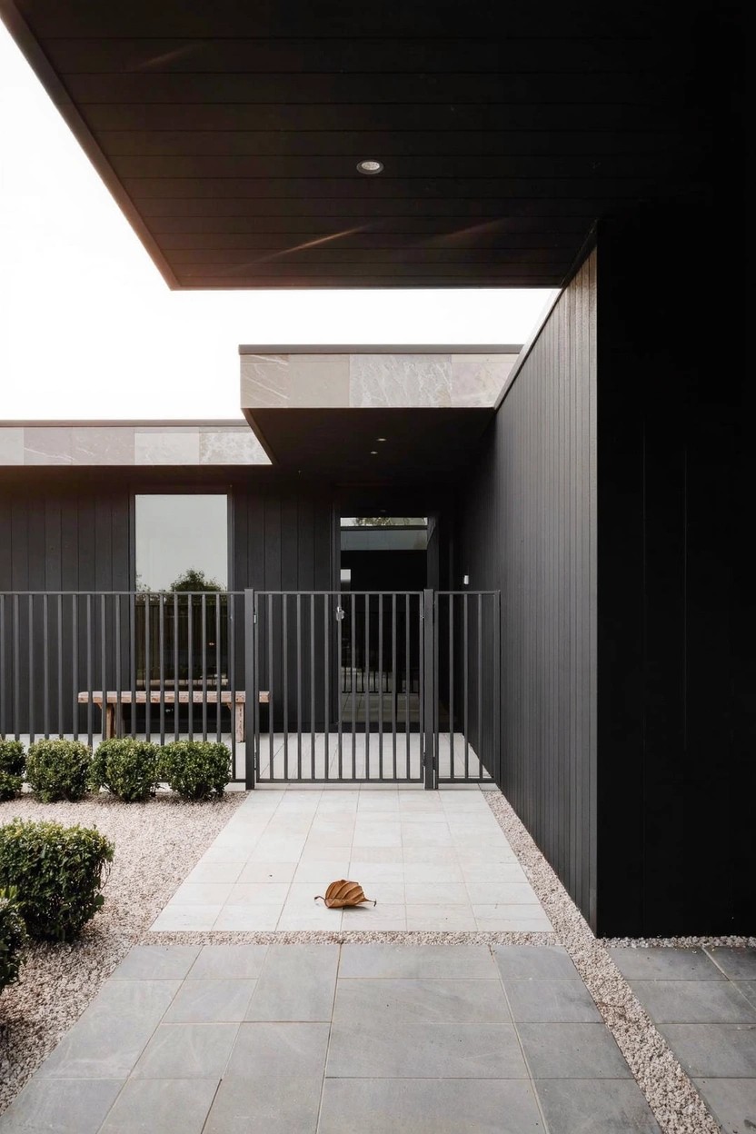 Modern one-story house exterior with black cladding, flat overhanging roof, black metal entry gate, concrete bench, gravel-bordered paver pathway, and low shrubs.