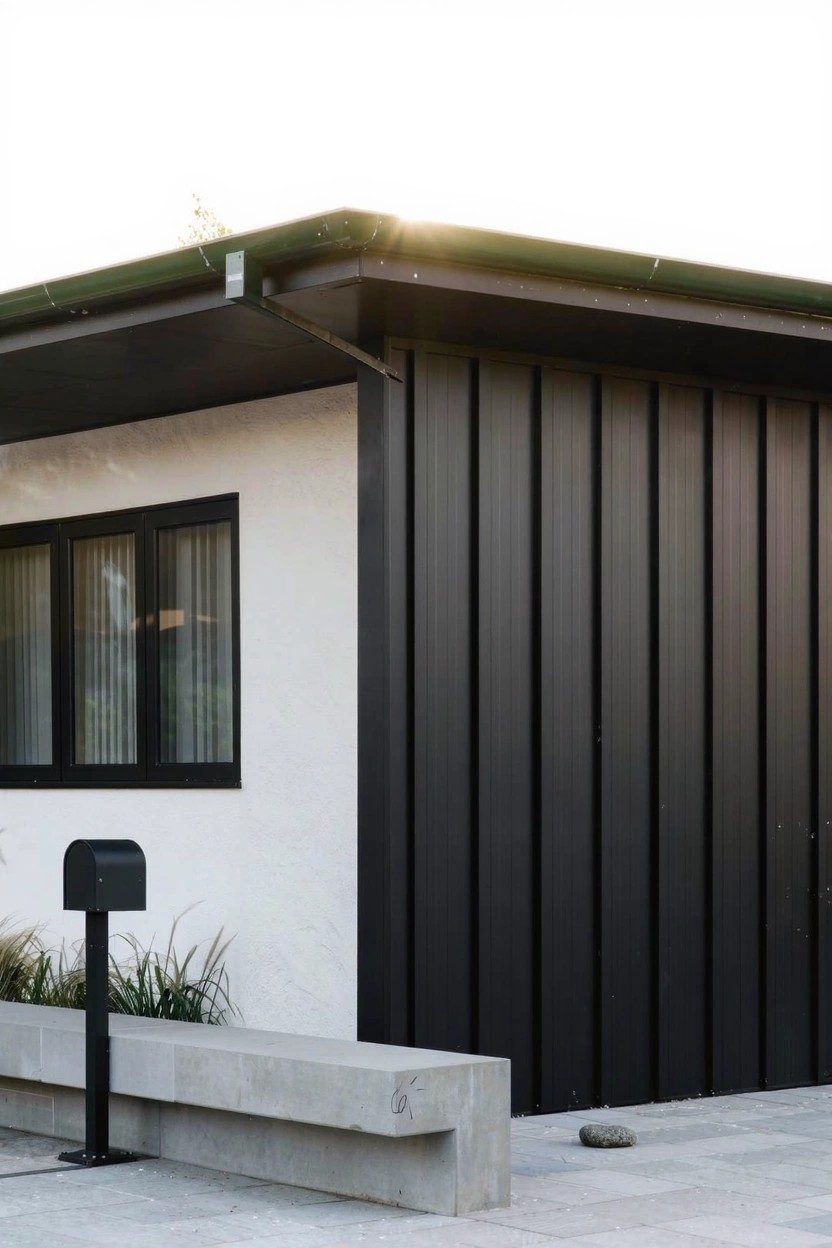 Corner view of a single-story modern house with white walls, black vertical siding panels, large triple-pane windows with black frames, metal mailbox post, concrete bench, pavers, ornamental grasses, and a stone nearby.
