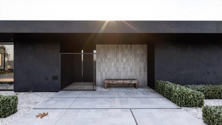 Modern one-story house exterior with black cladding, flat overhanging roof, black metal entry gate, concrete bench, gravel-bordered paver pathway, and low shrubs.
