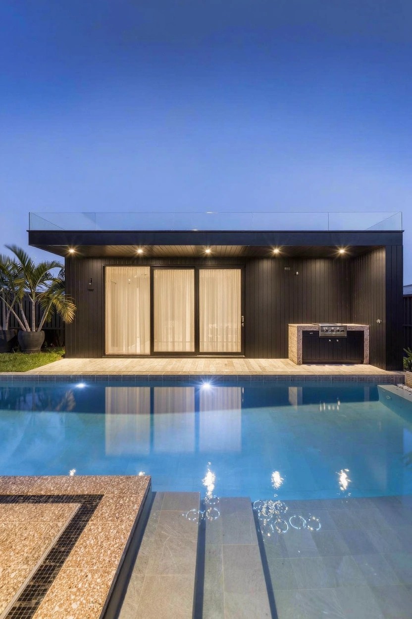 Modern black single-story cabana with glass sliding doors, sheer curtains, and adjacent outdoor kitchen cabinet next to a rectangular pool with decking and steps at dusk.