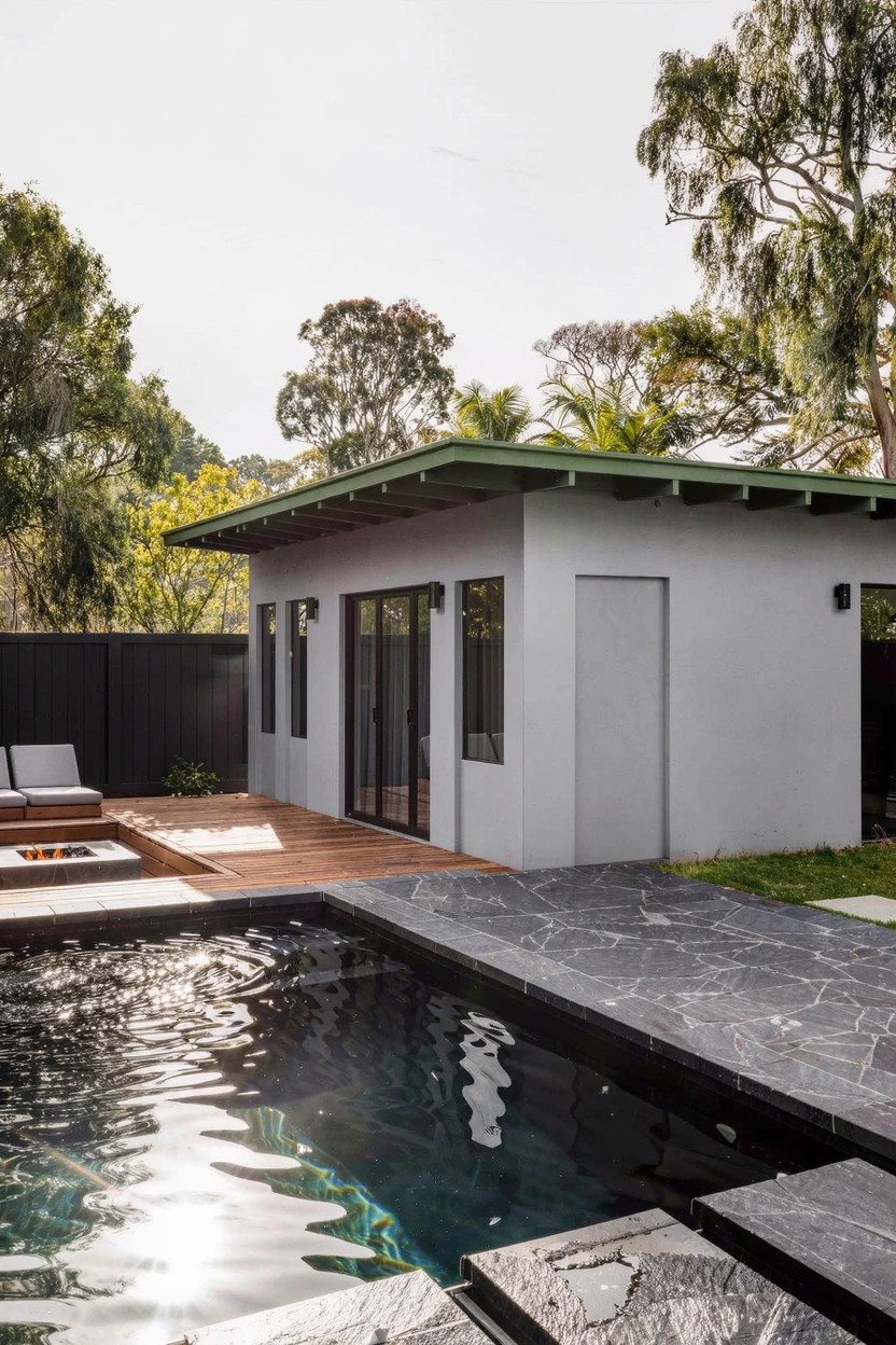 White stucco pool house with green metal roof and large glass doors facing a wooden deck with loungers and fire pit next to a pool edged in black stone, trees and fence in background.