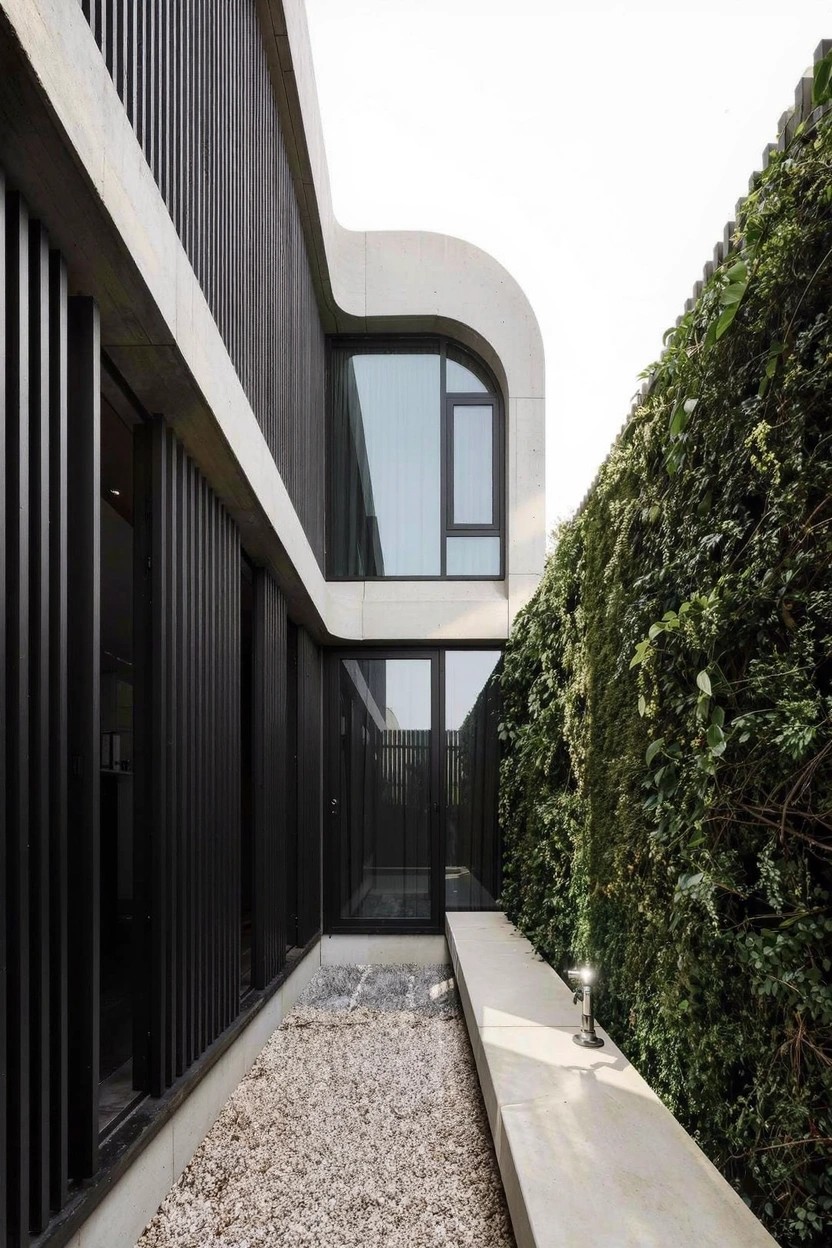 Modern one-story house exterior with black vertical slats on walls, curved white concrete section, glass windows and entry door, gravel path with bench and lamp, next to tall green ivy-covered wall.