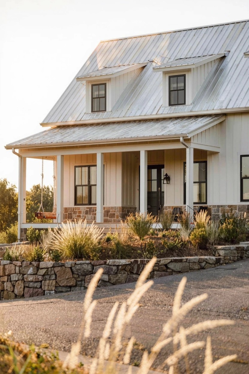 White board-and-batten house with standing seam metal roof, covered front porch supported by posts, black-framed windows and door, stone retaining wall at base, potted swing on porch, ornamental grasses, and gravel driveway at sunset.