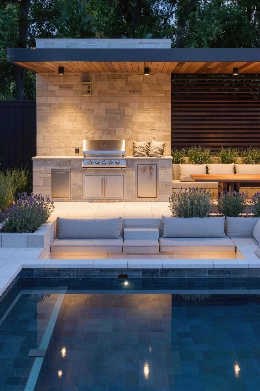 Backyard patio with a stone-built outdoor kitchen including grill and cabinets, built-in cushioned seating around a rectangular pool, wooden slat privacy screens, potted lavender plants, and dusk lighting.
