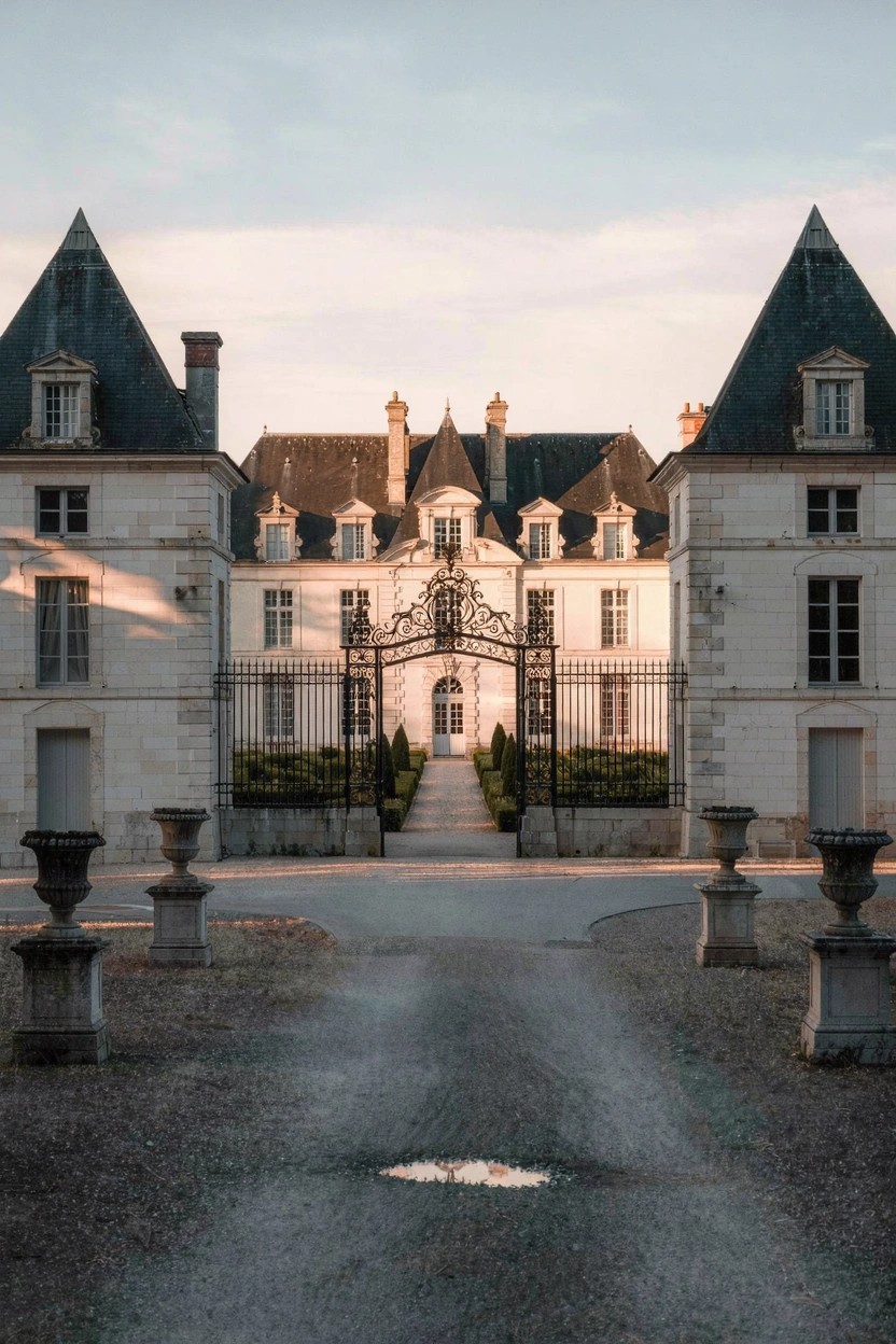 White stone French chateau with symmetrical wings, steep dark slate roofs, central wrought-iron gates, stone urns, and gravel driveway.