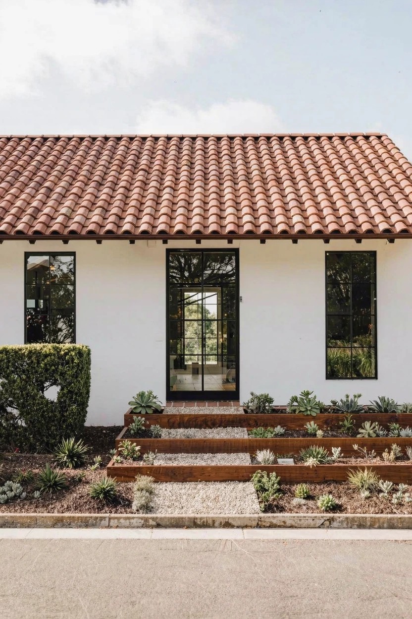 White stucco single-story house with red tile roof, black metal-framed glass front door and side windows, gravel steps leading up to entry flanked by raised succulent plantings and trimmed hedges.