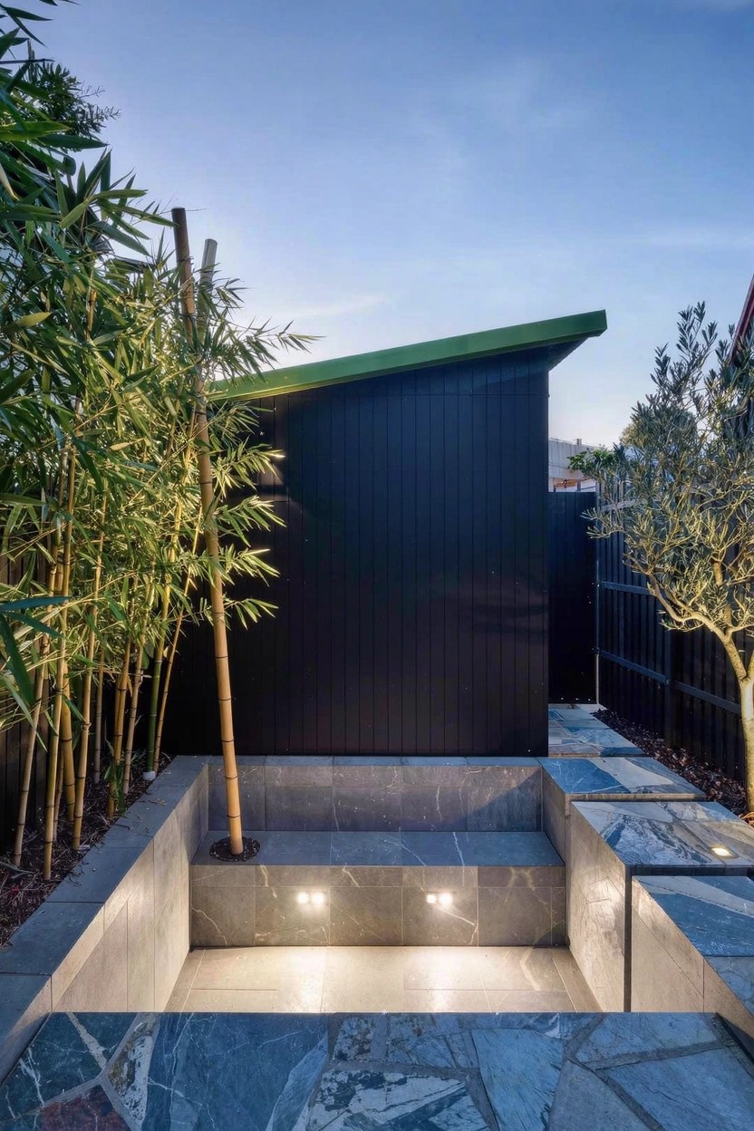 Backyard view showing a black timber wall with green roof, surrounded by bamboo and an olive tree, next to a sunken rectangular grey stone platform with underlighting and a stone tile pathway.