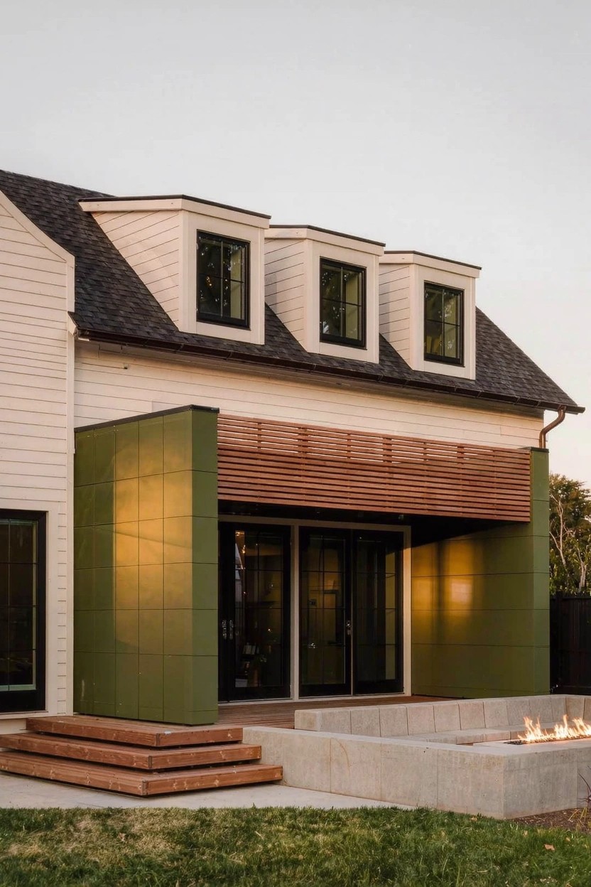 White-sided house with green vertical panel wall, wood slats over glass entry doors, dormer windows on roof, steps to concrete patio with linear fire pit and grass yard.
