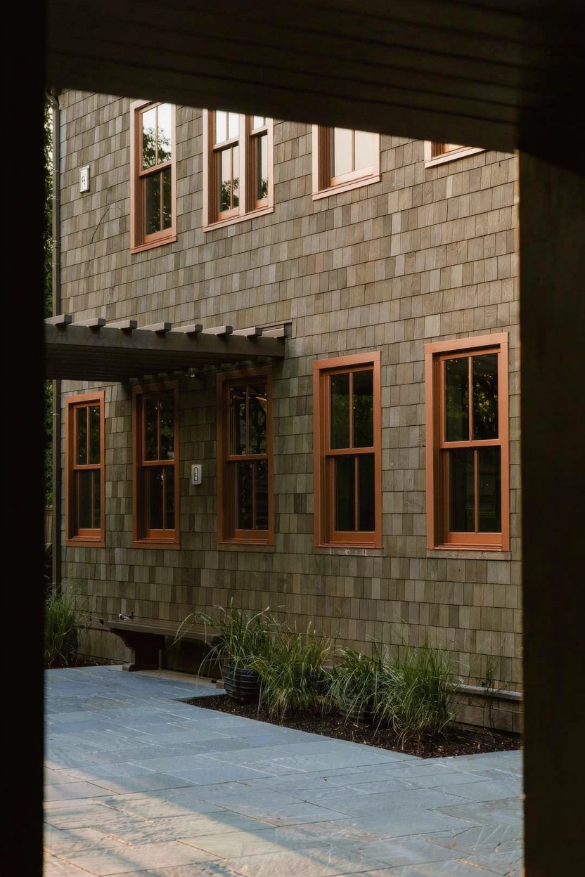 Shingled house exterior in gray with orange-brown window frames, viewed from a covered entry with stone pavers and potted grasses nearby.