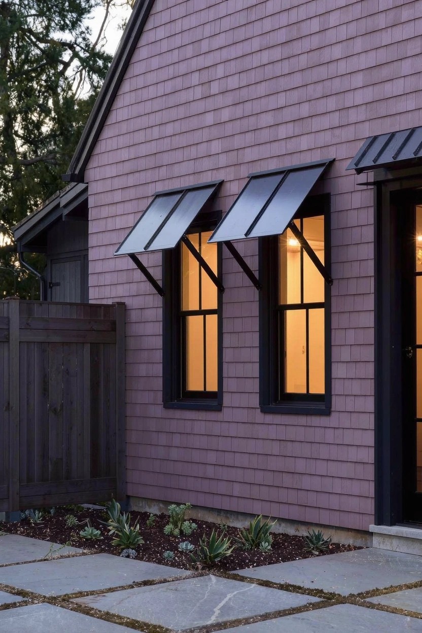 House exterior with mauve shingle siding, black-framed windows under metal awnings, dark wood entry door, wooden fence, succulent plants along stone paver path, and trees in background at dusk.