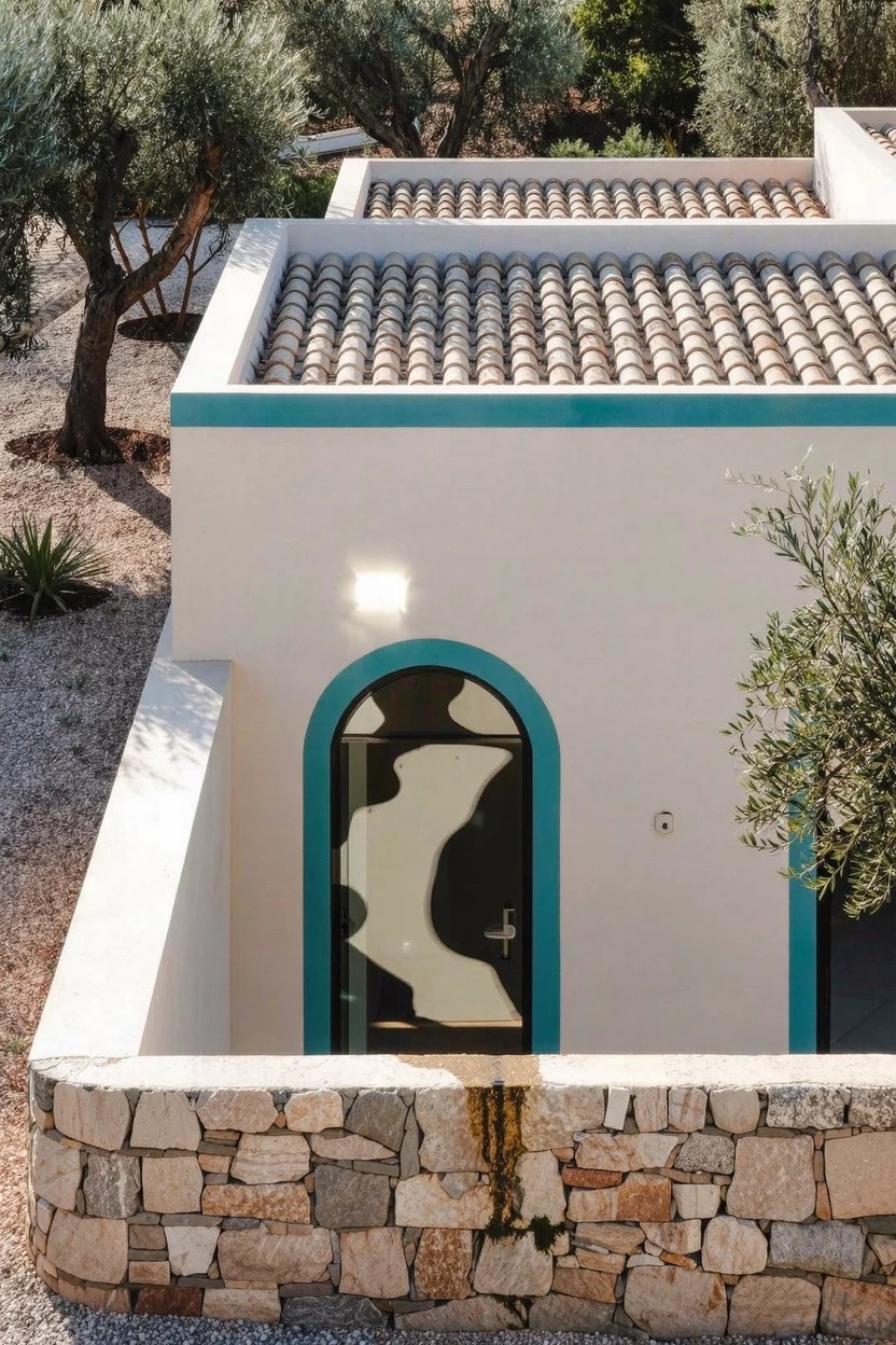 White stucco house exterior with teal trim around the arched black door and roof edges, terracotta tile roof, olive trees, stone retaining wall, and gravel ground.